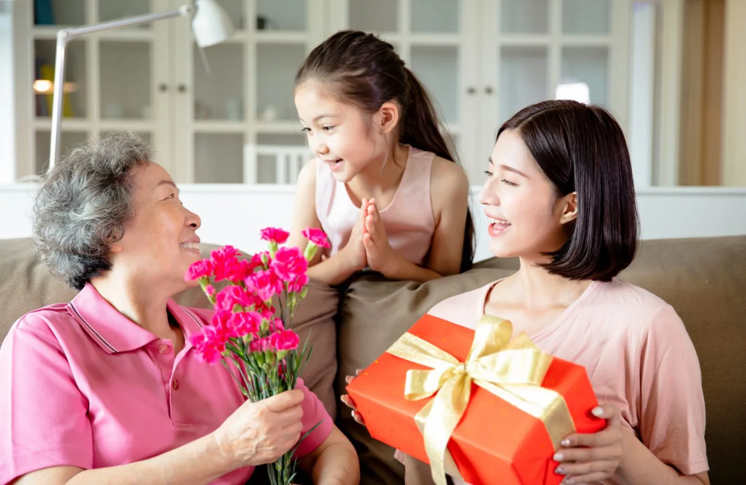 Ba người đang ngồi trên ghế, trao nhau hoa và hộp quà, với bối cảnh phòng khách sáng sủa.
