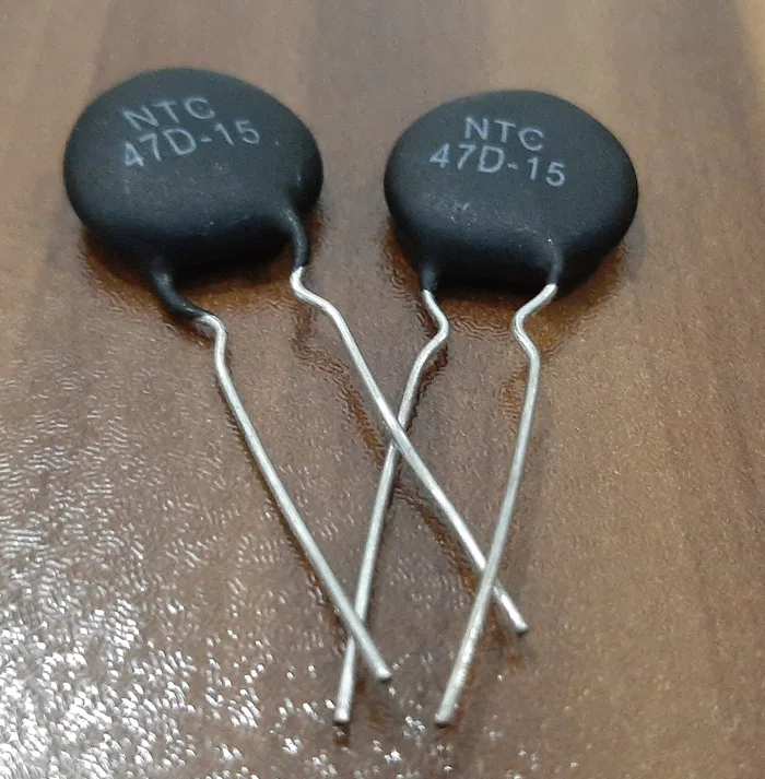 Ảnh chụp hai nhiệt điện trở NTC hình đĩa màu đen in chữ NTC 47D-15, hai chân dài, đặt trên mặt bàn gỗ