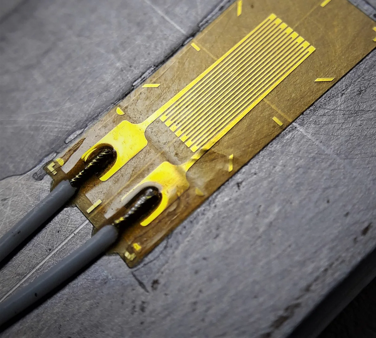 Strain gauge lá kim loại màu vàng đồng dán trên bề mặt kim loại xám, lưới mảnh song song, hai chỗ hàn lớn ở một đầu nối với hai dây bọc xám