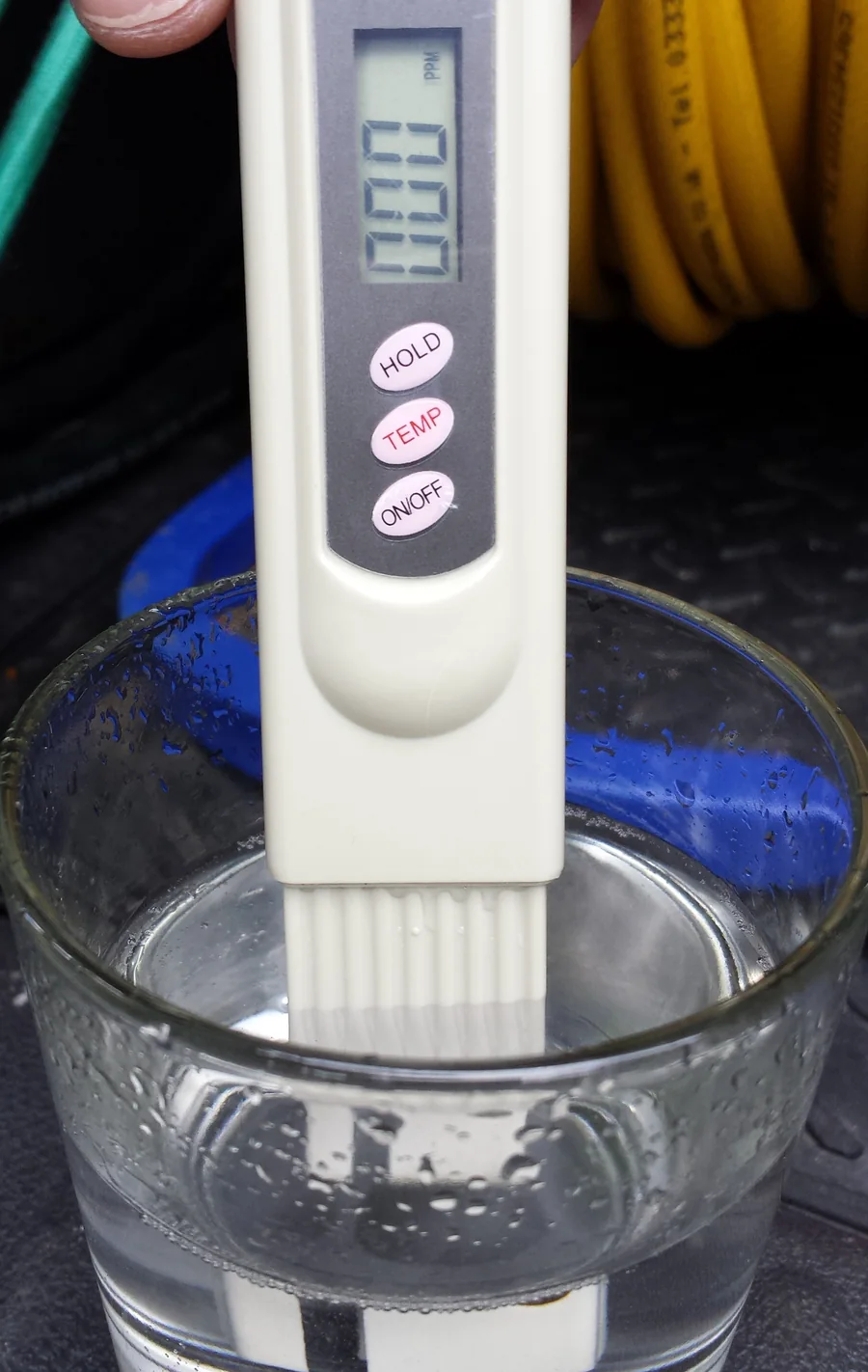 Bút đo TDS màu trắng nhúng đầu đo vào cốc nước thủy tinh, màn hình LCD hiện số, ba nút HOLD, TEMP, ON/OFF trên thân bút