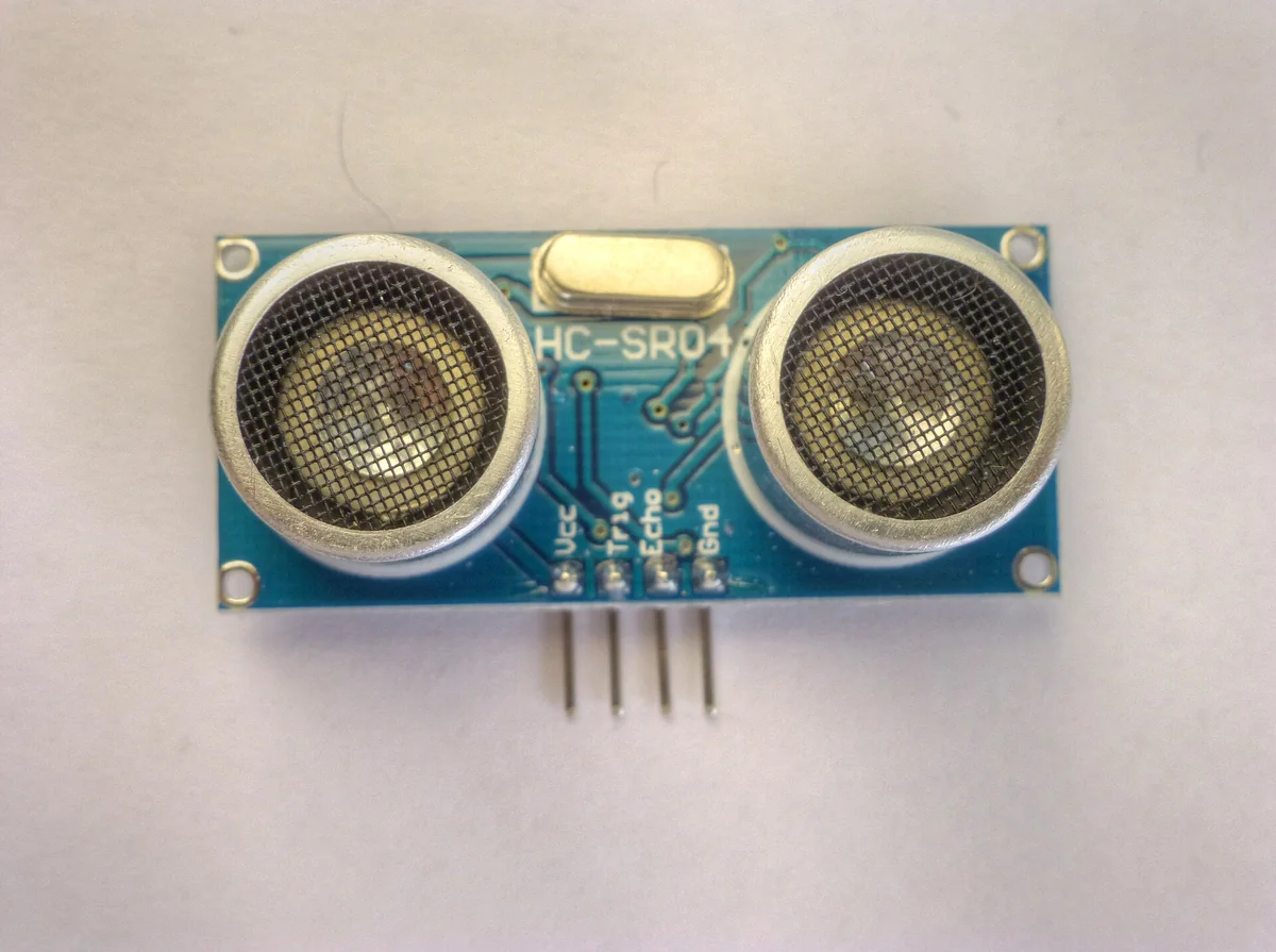 Module cảm biến siêu âm HC-SR04 màu xanh với hai đầu dò tròn có lưới kim loại, một thạch anh ở giữa và bốn chân Vcc, Trig, Echo, Gnd