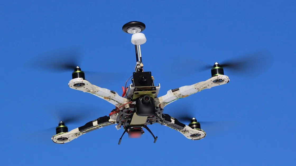 Một quadcopter tự lắp ráp đang bay trên nền trời xanh: bốn tay đòn màu trắng, bốn động cơ ở đầu tay đòn, ở giữa là camera, mạch điện và cột ăng-ten GPS