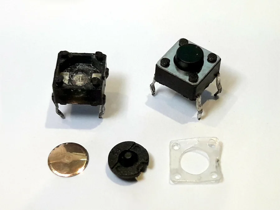 Ảnh chụp một nút nhấn tactile còn nguyên bên phải và một nút đã tháo rời bên trái: vỏ đen có bốn chân, miếng vòm kim loại tròn, nút bấm đen và nắp nhựa trong