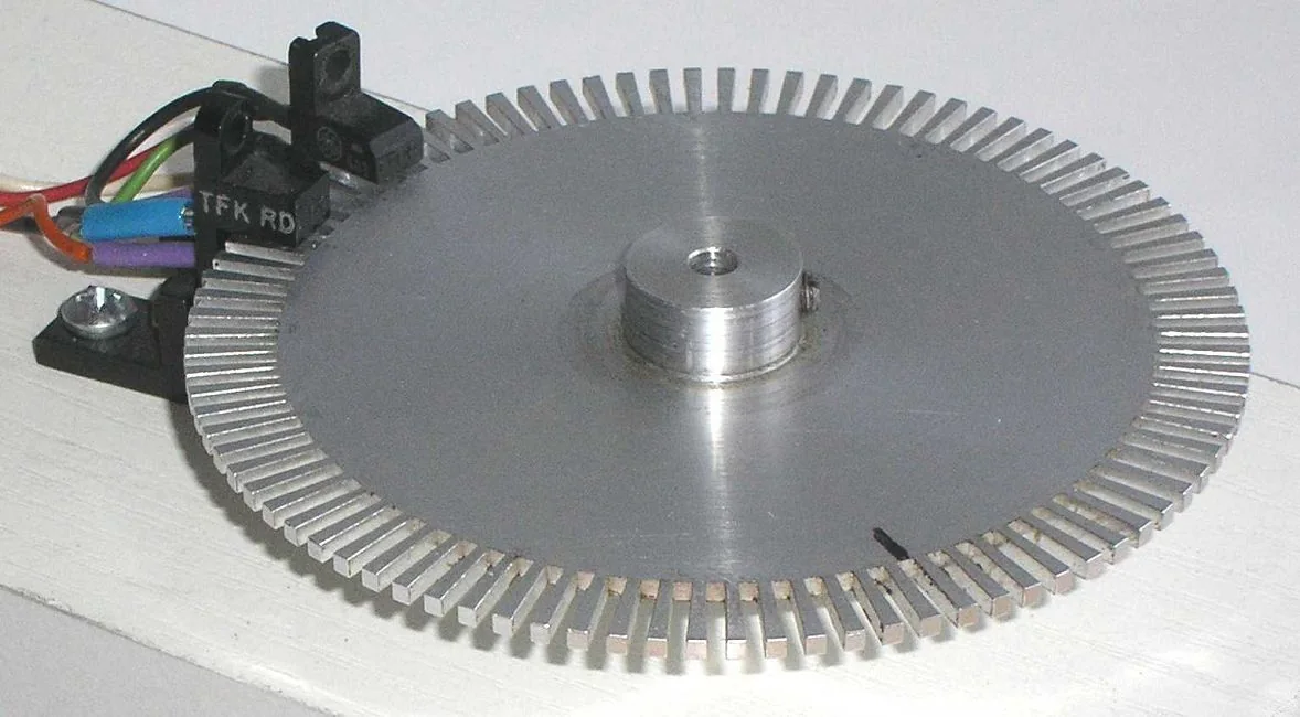 Mô hình encoder quang: đĩa nhôm tròn có nhiều rãnh ở mép, một cảm biến quang chữ U ôm lấy mép đĩa