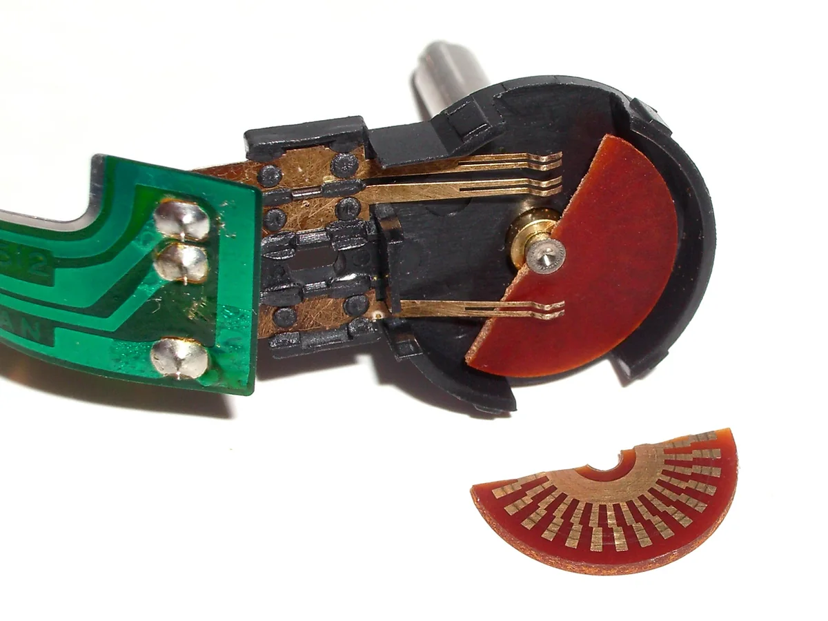 Encoder cơ khí tháo ra từ một con chuột máy tính cũ: đĩa nhựa màu đỏ có các vạch đồng dẫn điện, ba lá đồng tiếp điểm trên đế nhựa đen gắn vào mạch in