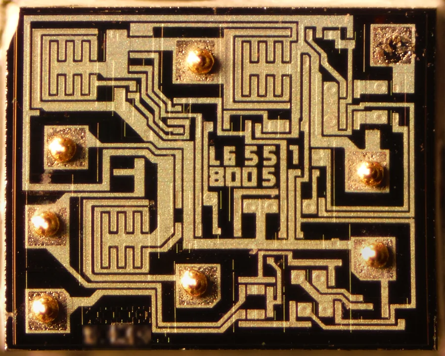 Ảnh chụp hiển vi miếng silicon bên trong IC NE555: các đường kim loại màu vàng nhạt trên nền đen, các vùng hình răng lược là transistor, tám điểm hàn tròn màu vàng ở mép nơi dây nối đi ra tám chân, ở giữa có dòng chữ L655 8005