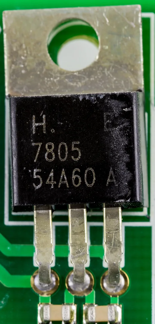 Ảnh chụp cận cảnh IC ổn áp 7805 vỏ TO-220 hàn trên mạch in màu xanh lá: tấm tản nhiệt kim loại có lỗ bắt vít ở trên, thân nhựa đen in chữ 7805, ba chân kim loại hàn xuống mạch, bên dưới là hai tụ dán nhỏ