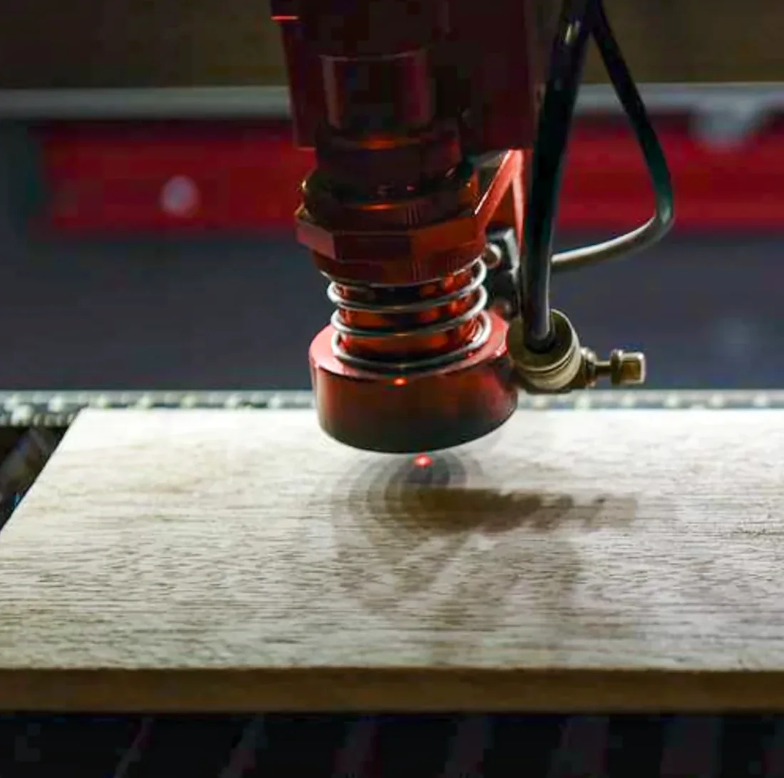 Đầu cắt laser màu đỏ có lò xo đang chiếu điểm sáng nhỏ xuống tấm gỗ, vệt cháy xém mờ quanh điểm khắc