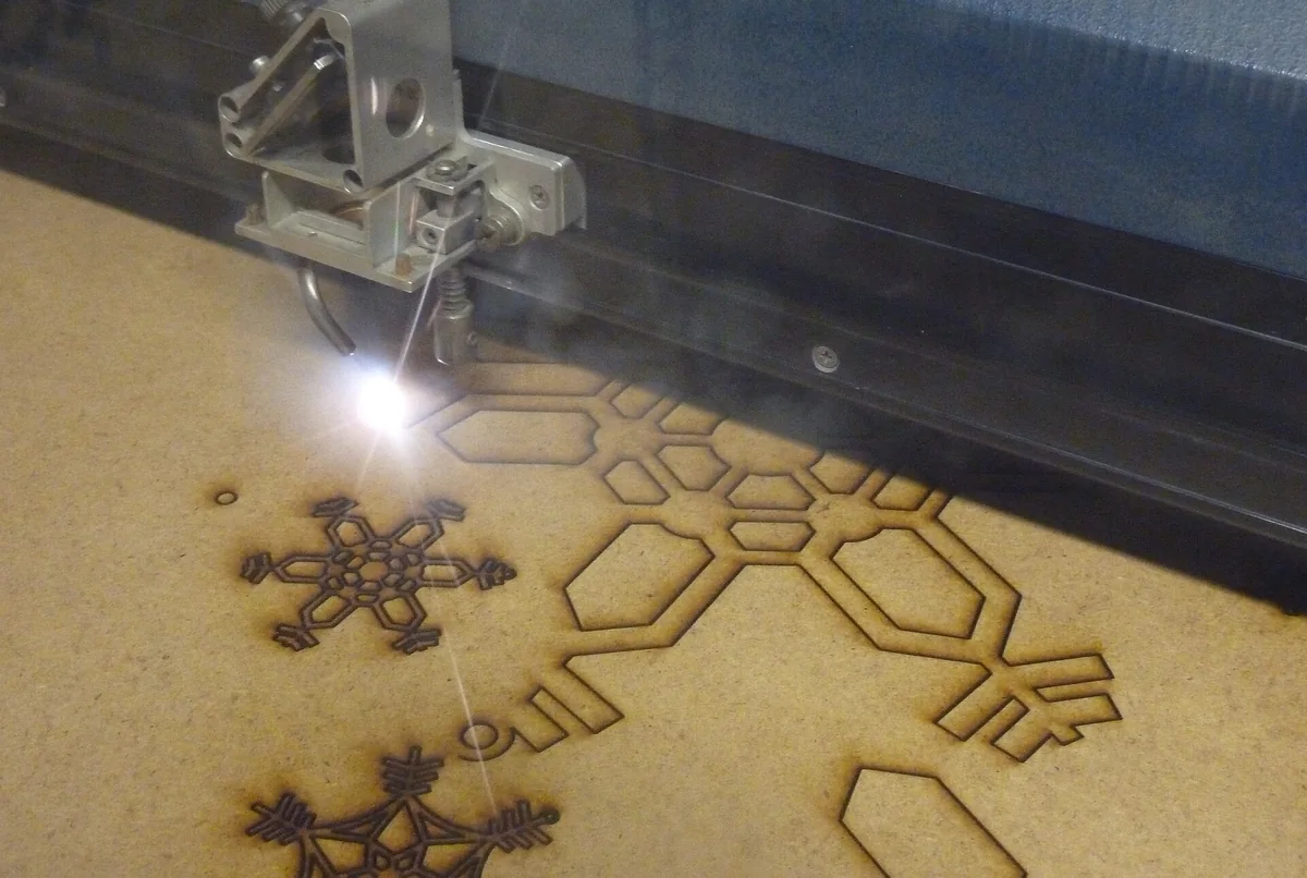 Đầu laser của máy cắt đang cắt hoa văn bông tuyết trên tấm gỗ MDF, điểm sáng chói tại chỗ cắt, các đường cắt màu nâu sẫm