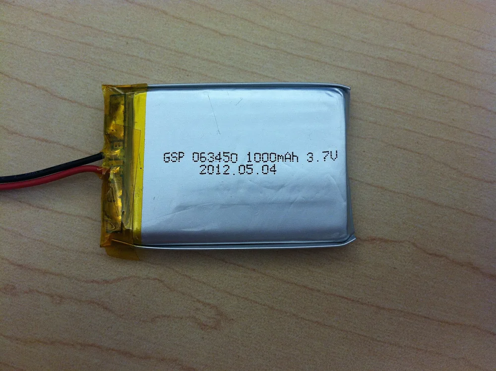Một cell pin LiPo dạng túi bạc, dán băng keo vàng ở đầu có mạch bảo vệ, hai dây đỏ đen, in dòng chữ 063450 1000mAh 3.7V