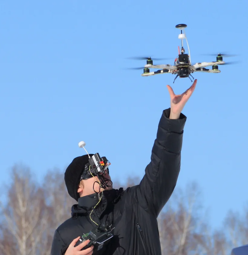 Người lái đeo kính FPV tự chế có ăng-ten nhỏ trên đầu, một tay cầm tay điều khiển, tay kia vừa thả một quadcopter lên khỏi bàn tay trên nền trời xanh