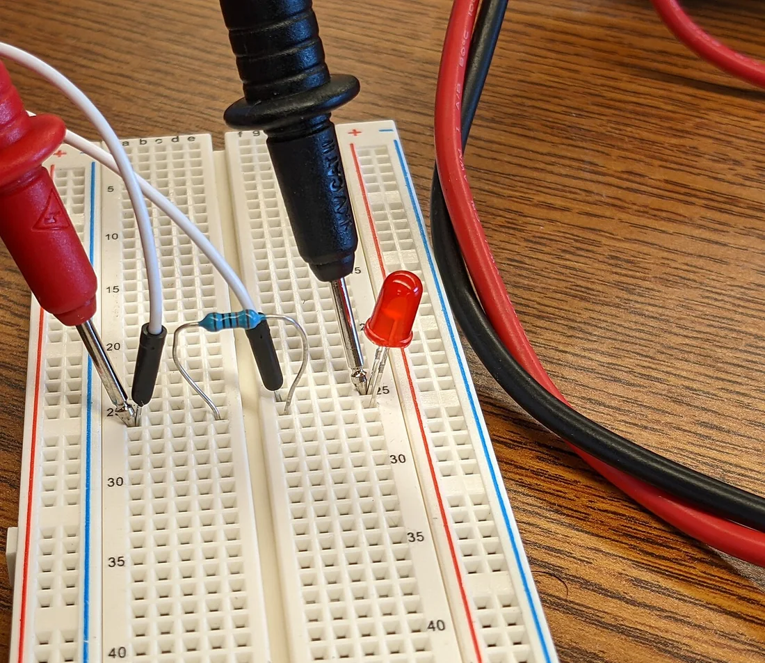 Ảnh chụp hai que đo đỏ đen của đồng hồ vạn năng chạm vào hai hàng lỗ của breadboard trắng, trên breadboard có một điện trở vòng màu, dây cắm trắng và một LED đỏ 5 mm