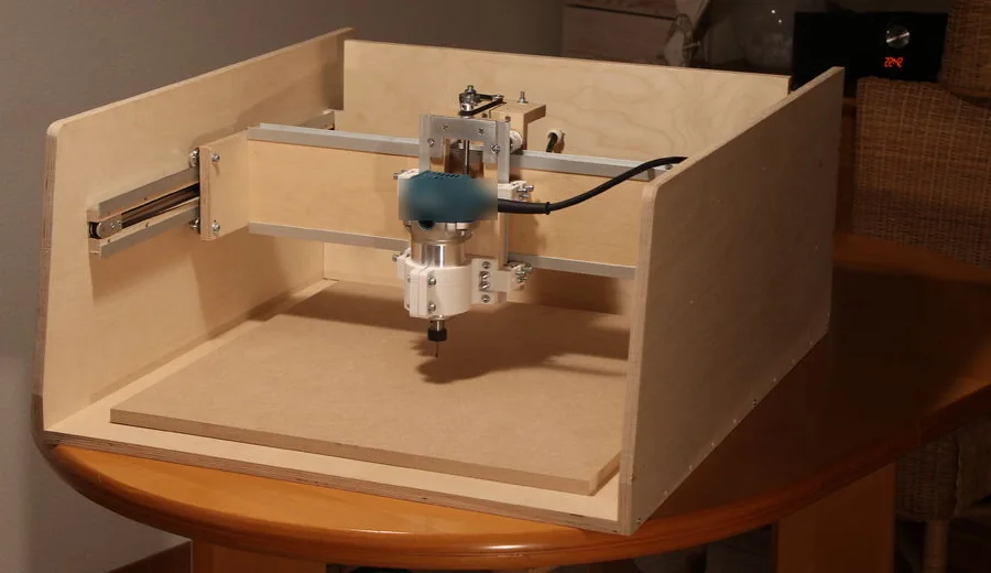 Máy CNC tự chế khung gỗ dán đặt trên bàn tròn: hai vách gỗ hai bên, ray trượt bên trái, dầm ngang bằng gỗ và thanh nhôm, máy phay cầm tay kẹp trên bàn trượt Z, mũi phay chĩa xuống tấm lót MDF