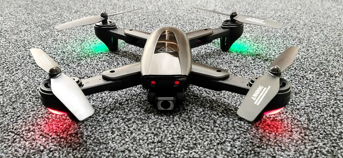 Flycam gập cỡ nhỏ đặt trên nền xám, bốn tay đòn đã mở, cánh quạt và camera phía trước